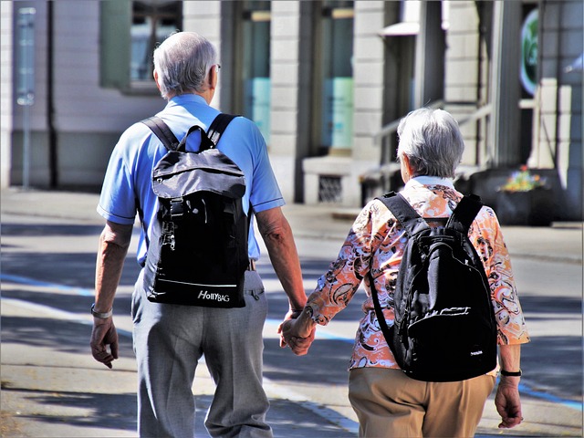 An elderly man and woman seen from behind walking hand in hand along a sunny urban sidewalk, each carrying a backpack.